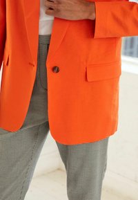 Blazer arancione brillante con un solo bottone, abbinato a pantaloni a scacchi bianco e nero in stile houndstooth. Texture liscia, design strutturato.