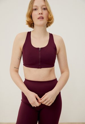 Γυναίκα με κοντά ξανθά μαλλιά, φοράει μπρούντζινο αθλητικό σουτιέν με φερμουάρ μπροστά και αντίστοιχα κολάν με ψηλή μέση, στέκεται μπροστά σε απλό φόντο.