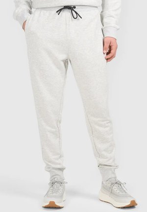 Jasnoszare spodnie dresowe ze sznurkiem w czarnym kolorze noszone przez stojącą osobę z rękami w kieszeniach, połączone z jasnoszarymi sneakersami z białymi podeszwami.