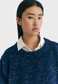 Pull en tricot bleu marine avec un col côtelé, porté sur une chemise blanche à col. Tissu texturé avec de légères speckles.