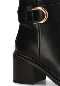 Bota de tobillo de cuero negro con punta redonda, un acento de hebilla en tono dorado en una correa ancha y un tacón cuadrado texturizado.
