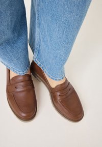 Personne portant un jean bleu clair et des mocassins en cuir marron se tenant sur un sol clair.