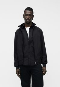 Μαύρος άνδρας που στέκεται φορώντας ένα μαύρο μπουφάν με φερμουάρ πάνω από ένα μαύρο πουκάμισο με κουμπιά και ένα λευκό εσώρουχο μπλουζάκι, μπροστά σε ένα απλό λευκό φόντο.