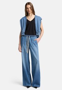 Jeansoutfit med ett ärmlöst ljusblått ytterlager och svart V-ringad topp, kombinerat med lösa blå joggerbyxor med snöre.