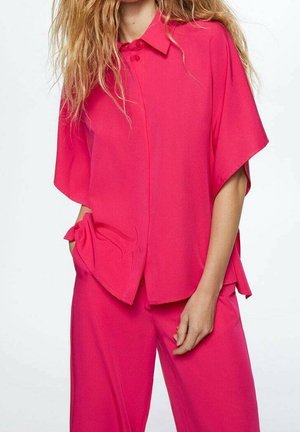 Woman wearing bright pink loose short-sleeve button-up shirt and matching wide-leg pants against a plain background.
