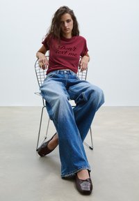 Bordeaux t-shirt med teksten "Don't text me", sammen med brede blå jeans og brune flade sko, siddende på en trådstol mod en ensfarvet baggrund.