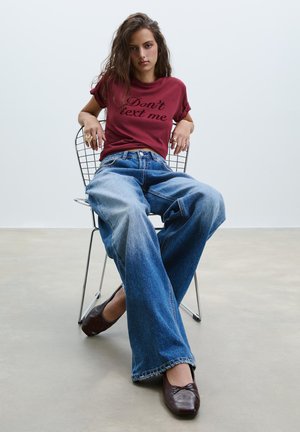 T-shirt bourgogne avec le texte "Ne m'écris pas", associé à un jean bleu à jambes larges et des ballerines marron, assis sur une chaise en fil contre un fond uni.