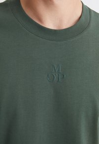 Grünes Baumwoll-T-Shirt mit Rundhalsausschnitt, das ein geprägtes "M O’P"-Logo auf der linken Brustseite aufweist. Glatte Textur, minimalistisches Design.