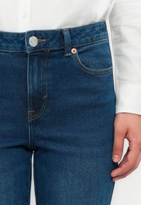 Nærbilde av blå denimjeans med knapp- og lomme-detaljer, brukt sammen med en hvit langermet skjorte, og en hånd hvilende ved siden.