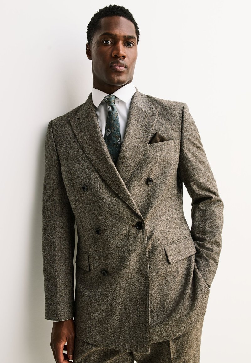 Hombre con un traje de tweed marrón cruzado, corbata con estampado floral y camisa blanca, de pie frente a un fondo blanco liso.
