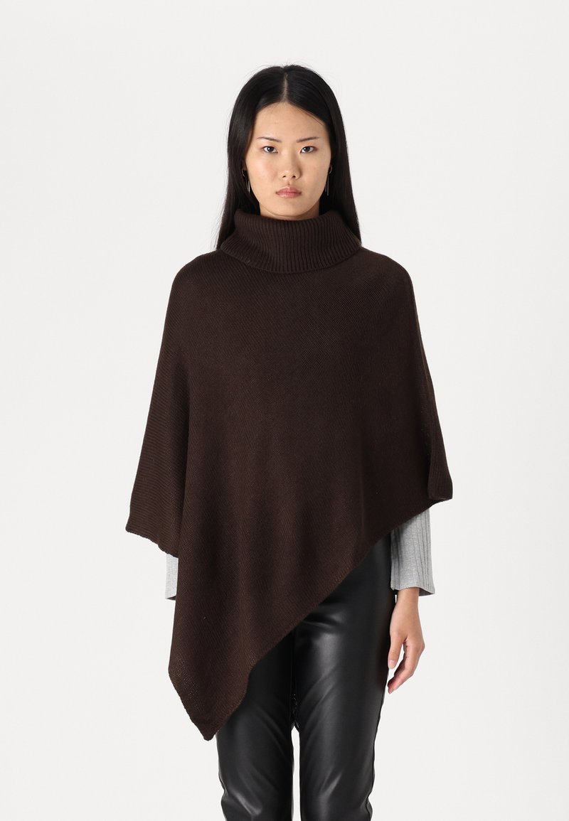 Brun strikket poncho med høy hals, asymmetrisk kant og ribbestrikket tekstur, drapert over en grå langermet topp og lærbukser.