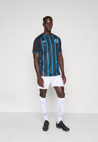 Homem em pé, vestindo uma camisola de futebol preta com riscas verticais azuis, calções brancos, meias até ao joelho brancas e chuteiras pretas, em fundo plain.