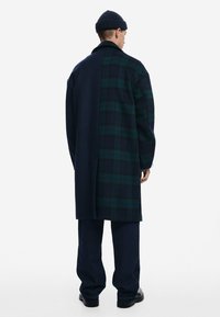 Long wool coat with a dark navy side and a green and navy plaid side. Finished with a collar, worn over dark denim pants and boots.