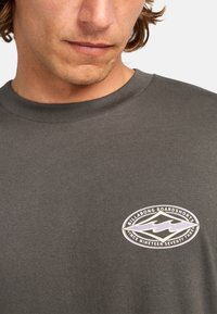 Gray cotton t-shirt featuring a round neckline and a logo patch on the left chest with a white and blue design detailing.