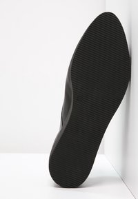 Chaussure noire avec une semelle en caoutchouc texturé, présentant des motifs striés et un design élégant. Le talon est légèrement surélevé, ce qui améliore la stabilité.