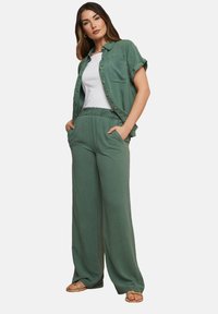 Camicia verde a maniche corte con bottoni sopra un top bianco, abbinata a pantaloni verdi larghi con vita elastica. Il modello è in piedi con le mani nelle tasche.