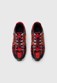 Melnas un sarkanas Nike sporta apavus ar režģa virsmas materiālu, atstarojošām svītrām, oranžiem Swoosh logo un melniem un sarkaniem auklām uz baltā fona.