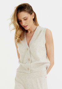 Cardigan sans manches beige en tricot doux, avec décolleté en V, bords côtelés et quatre boutons à l'avant. Détail subtil du logo près de l'ourlet.