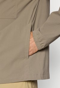 Cămașă de un maro deschis, cu un croi relaxat, având un buzunar aplicat, mansete elastice și detalii de cusătură vizibile. Material cu o textură moale și netedă.