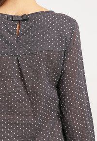 Blusa grigio scuro con piccoli motivi a rombi bianchi, apertura a occhiello sul retro e un piccolo dettaglio a fiocco sullo scollo. Tessuto leggero.