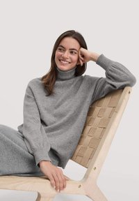 Grijze wollen coltrui met ribgebreide manchetten, gecombineerd met bijpassende grijze broek. Model zit op een licht houten stoel met een geweven zitting.