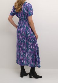 Robe longue bleu sarcelle avec un motif abstrait violet et blanc, manches courtes bouffantes, taille cintrée, portée avec des bottes noires montantes au genou.