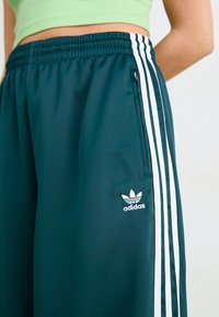 Tirkīza sporta bikses ar baltām sānu svītrām, elastīgu jostu un mazu ar rāvējslēdzēju aizveramu kabatu. Ietver iespiestu Adidas logotipu baltā krāsā.