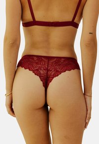String en dentelle bordeaux avec motif floral, bords scallopés et texture douce. Comprend des bretelles ajustables et un design à couverture minimale.