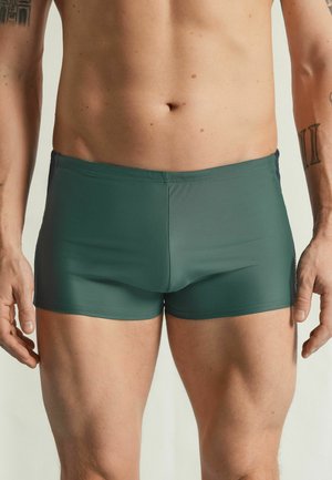 Męski tors i górna część nóg w ciemnozielonych kąpielówkach, z widocznymi tatuażami na obu ramionach na jednolitym jasnym tle.