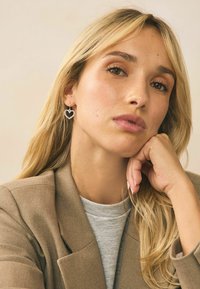 Orecchini a forma di cuore in argento con una finitura brillante, indossati da una persona con i capelli lunghi, in un blazer dai toni neutri sopra una camicia grigia.