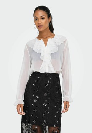 Blouse blanche transparente avec col volanté et patte de boutonnage, associée à une jupe noire ajustée ornée de motifs floraux en sequins.