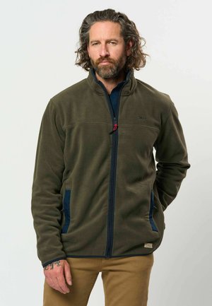 Hombre con barba y pelo rizado que lleva una chaqueta de forro polar verde oliva con cremallera azul marino y pantalones color beige, de pie sobre un fondo blanco.