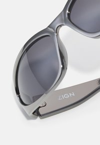 Černé oversized sluneční brýle s lesklým rámem, tmavě šedými čočkami a stříbrným logem ZIGN na spáncích. Hladký povrch a zakřivený design.