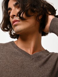 Maglione a V in cotone marrone con una texture morbida; il modello ha capelli ondulati di lunghezza spalla e un profilo facciale sottile.