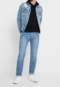 Denimjack met verwerkte details, zwarte hoodie eronder, lichtblauwe jeans en witte sneakers. De outfit heeft een casual, gelaagd uiterlijk.