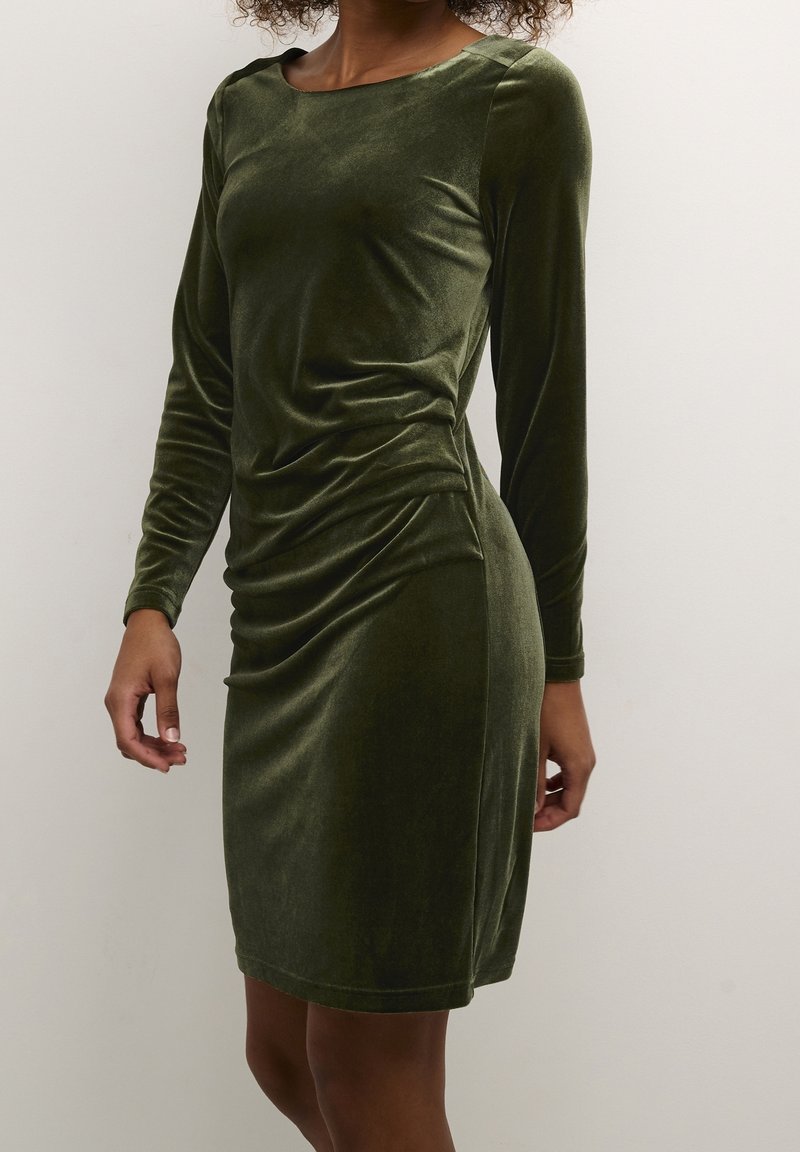 Robe en velours vert à manches longues, col bateau et détail froncé sur le côté, créant une silhouette ajustée. Texture lisse et douce.