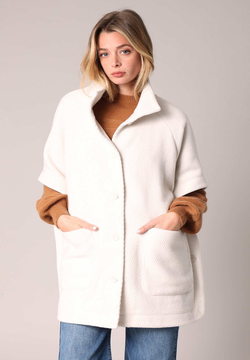 Weißer, übergroßer Wollmischmantel mit strukturierter Oberfläche, großen aufgesetzten Taschen und Stehkragen, über einem braunen Pullover getragen.