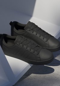 Paire de baskets basses noires avec lacets et bandes latérales discrètes, positionnées sur une plateforme blanche inclinée projetant des ombres sur un sol en béton.