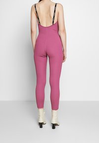 Kvinna som bär en ärmlös, rygglös rosa jumpsuit med svarta "Just Do It"-remmar och vita ankelstövlar, stående mot en vit bakgrund.