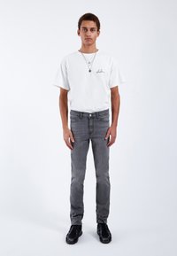 Camiseta blanca de manga corta con firma negra, combinada con jeans ajustados grises y zapatillas deportivas negras. Diseño simple y minimalista.