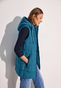 Teal pufferväst med huva, med quiltat mönster, dragkedjefront och sidofickor. Bärs över en mörk långärmad topp och blå jeans.