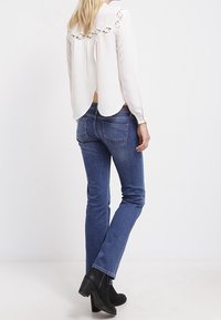 Blouse blanche avec des détails en dentelle au dos, associée à un jean bleu. Des bottes noires à la cheville complètent la tenue. Tissu lisse, coupe décontractée.