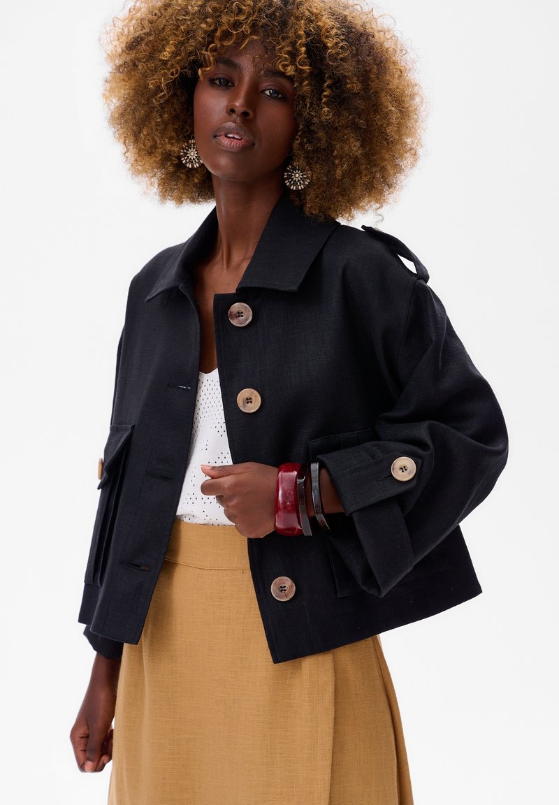 Czarna krótka kurtka z dużymi guzikami, dopasowany fason i kieszenie naszywane. Założona na białą bluzkę i beżową spódnicę z fakturowanej tkaniny.