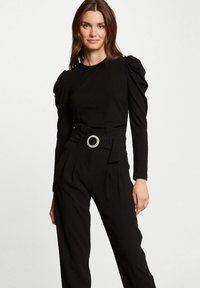Zwart geribbeld langmouwen top met opbollende schouders, gecombineerd met hoge zwarte broeken met een brede riem en een ronde zilveren gesp.