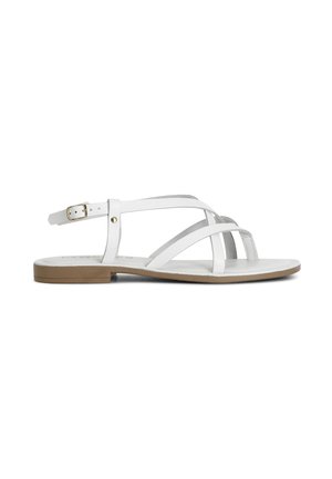 Hvid flad sandal med tynde stropper, der krydser tæerne og ankelrem med spænde, med lys brun sål og minimal hæl.