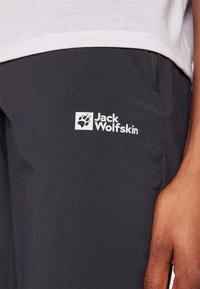 Mörkgrå utomhusbyxor med en slät textur, har en vit Jack Wolfskin-logotyp på sidan och en detalj med dragkedjeficka.