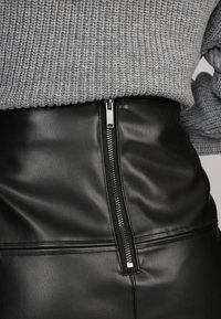 Jupe en simili cuir noir avec une fermeture éclair en argent visible et des détails de couture, associée à un pull en maille grise texturée au-dessus.