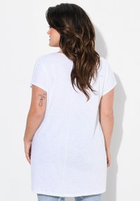 T-shirt blanc à manches courtes avec une coupe décontractée, doté d'un ourlet droit et d'une texture subtile. Un tatouage floral est visible sur le bras.