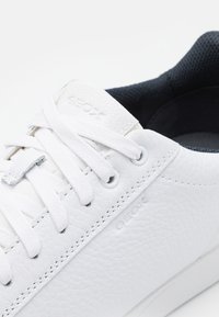 Primo piano di una sneaker in pelle bianca testurizzata con lacci bianchi, fodera interna nera e nome del marchio Geox impresso sulla linguetta e sul lato.