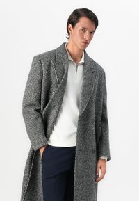 Mann in einem langen grauen gemusterten Übermantel, weißem Reißverschluss-Sweater und dunklen Hosen, steht mit einer Hand in der Tasche vor einem weißen Hintergrund.
